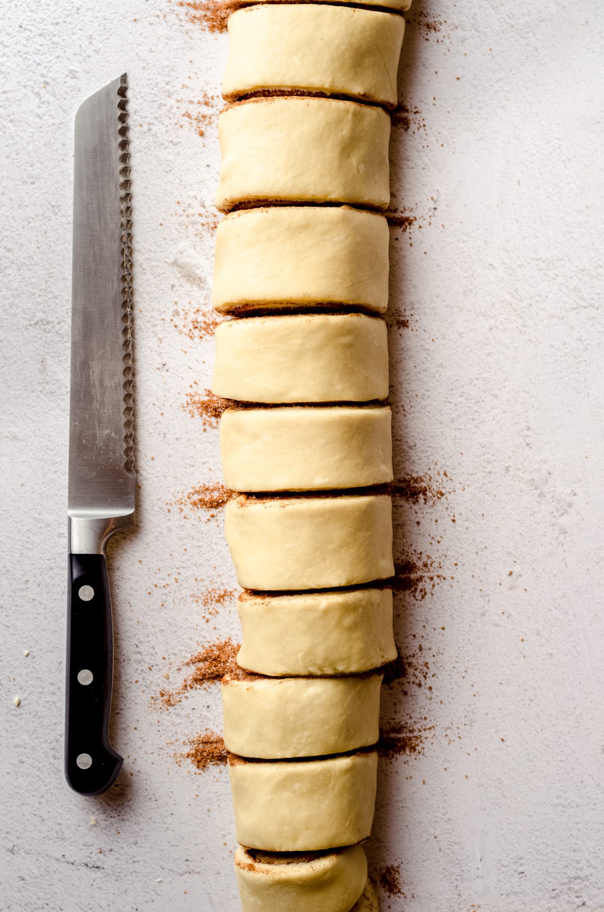 An aerial photo of a log of cinnamon roll dough sliced into portions with a serrated knife next to it.