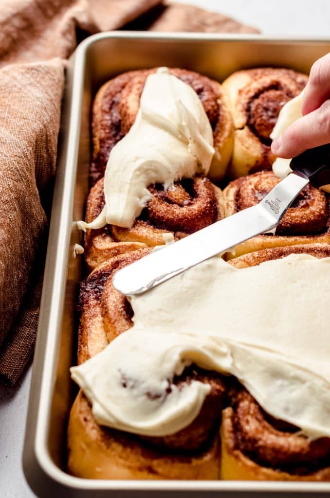 Someone using a small offset spatula to spread cream cheese frosting onto warm homemade cinnamon rolls.