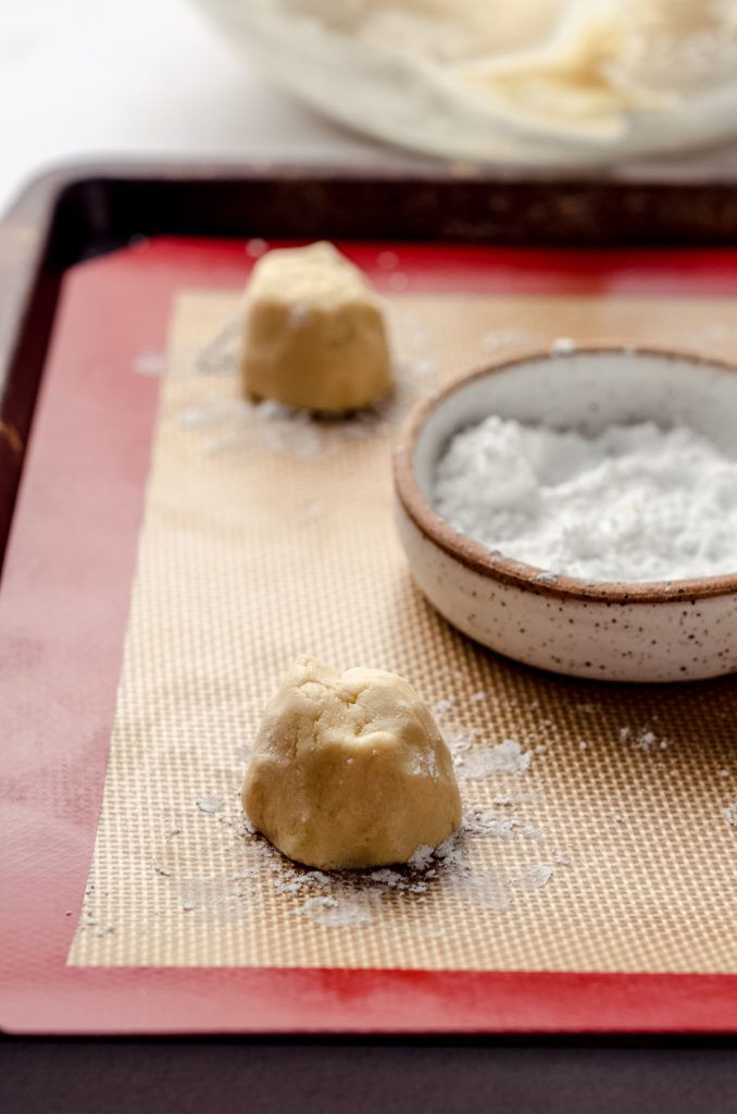 sugar cookie balls of dough on a baking sheet with a dish of powdered sugar