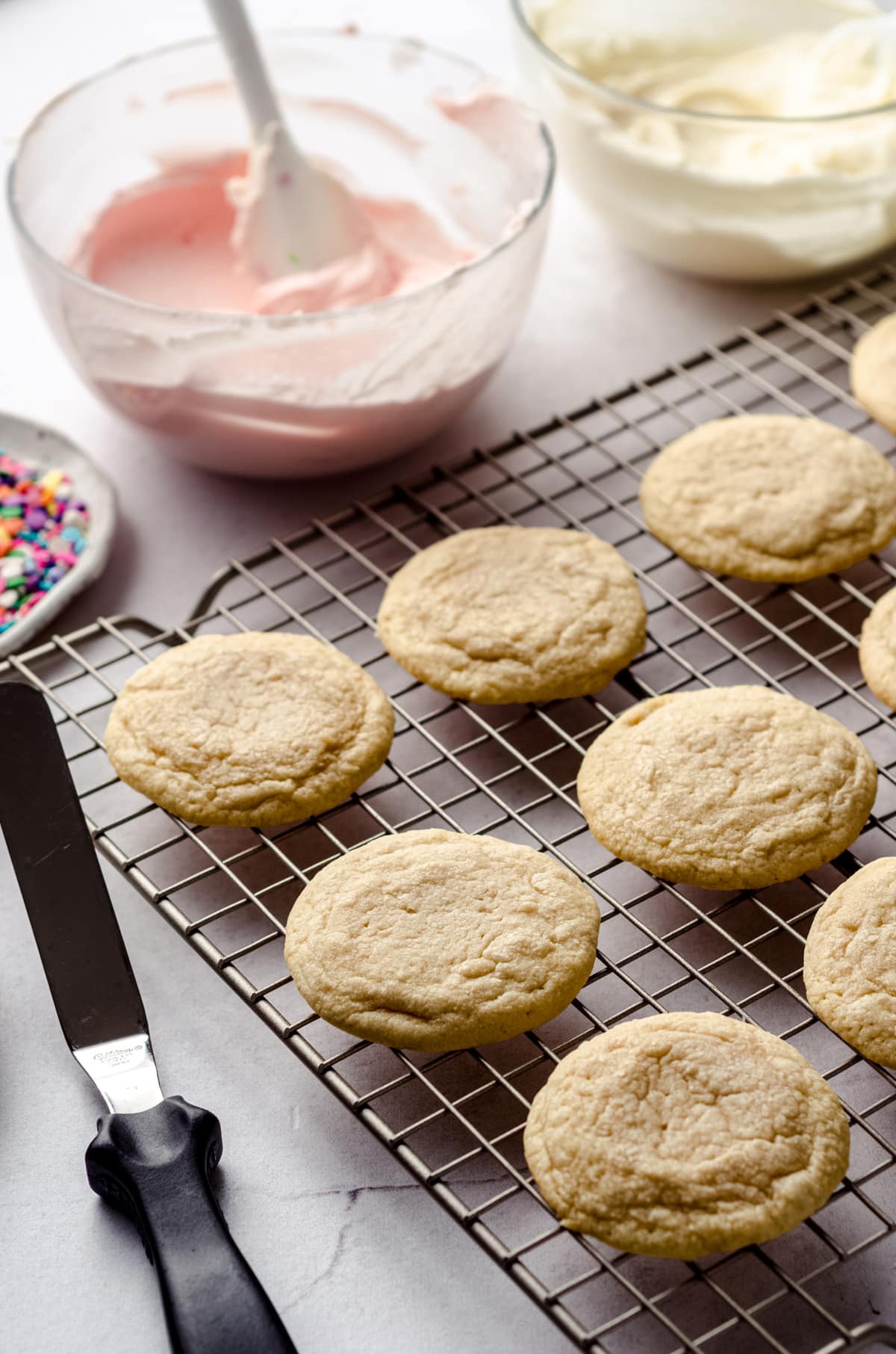sugar cookies on a cooling rack with bowls of frosting and tools for decorating
