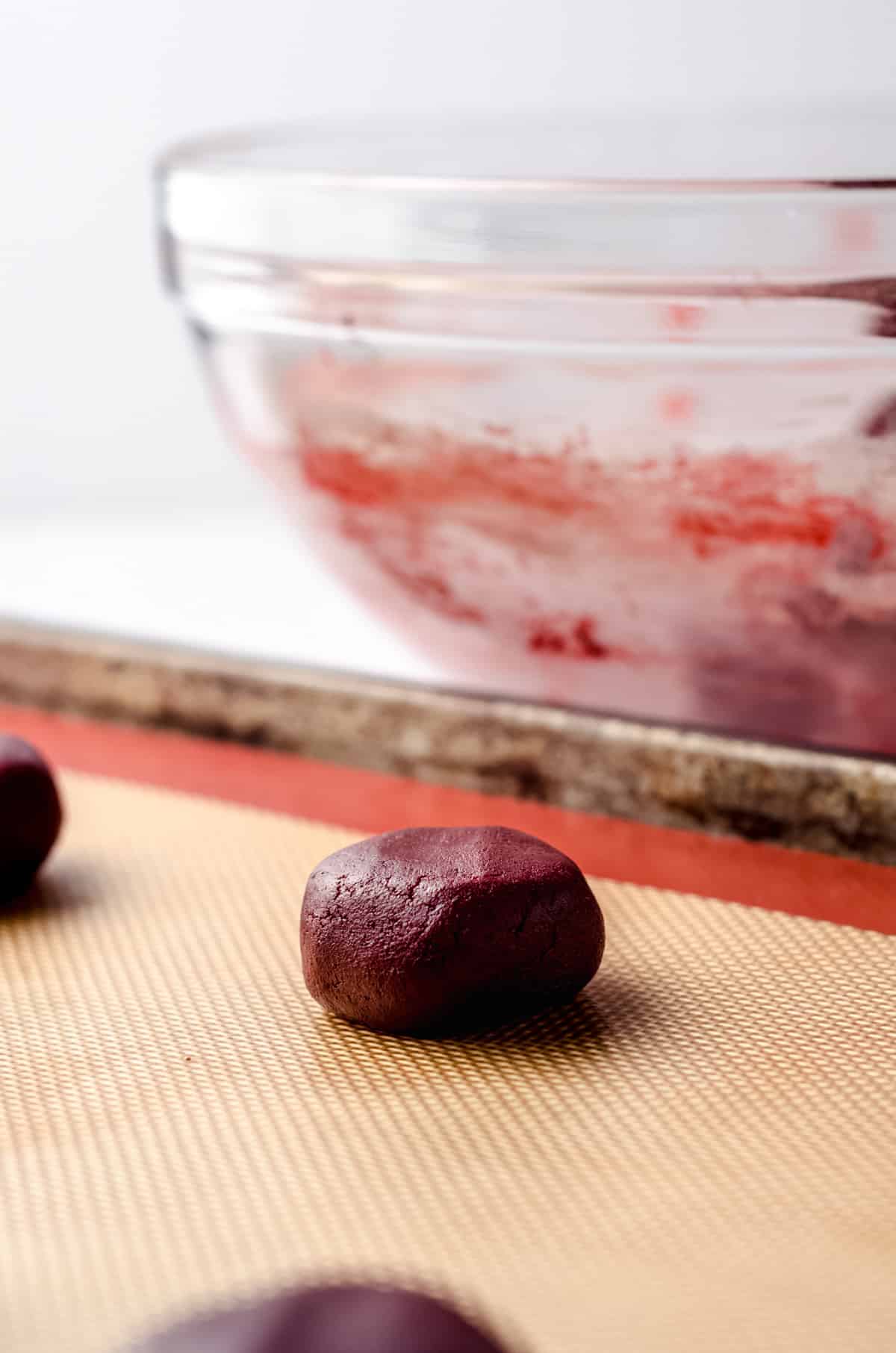 A ball of red velvet cookie dough is placed on a silicone mat in a baking tray. The ball of cookie dough is about the size of a golf ball. The glass bowl containing leftover dough is in the background.