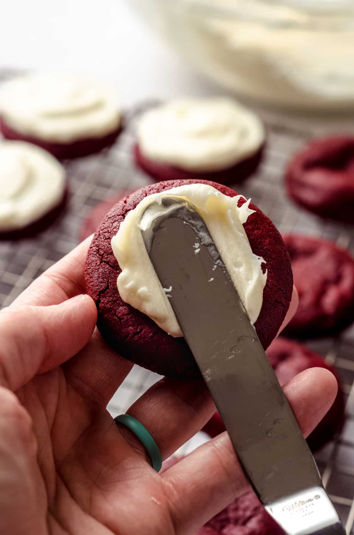 Someone is holding a red velvet cookie and, using a knife, is smearing it with cream cheese frosting.