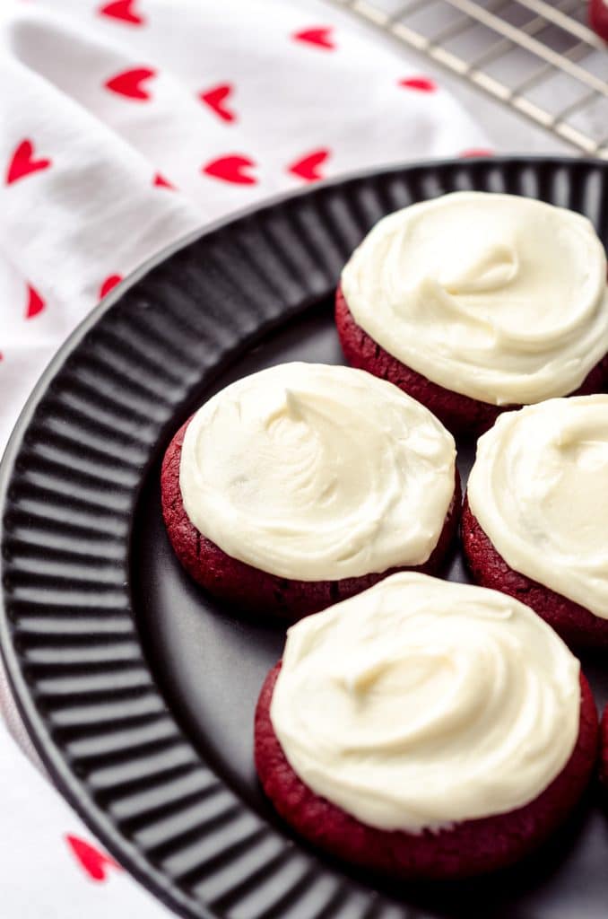 Half of a black plate can be seen. On top of this plate are red velvet cookies, topped with white cream cheese frosting.
