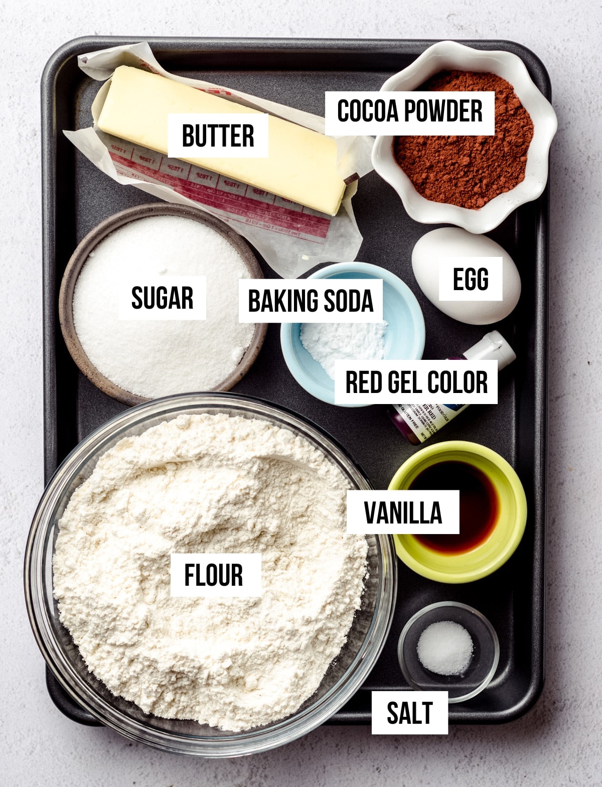 All the ingredients required for this recipe. There is a stick of butter, cocoa powder, a bowl of white sugar, a small bowl of baking soda, an egg, some red food gel color, vanilla, salt and a large glass bowl of flour.