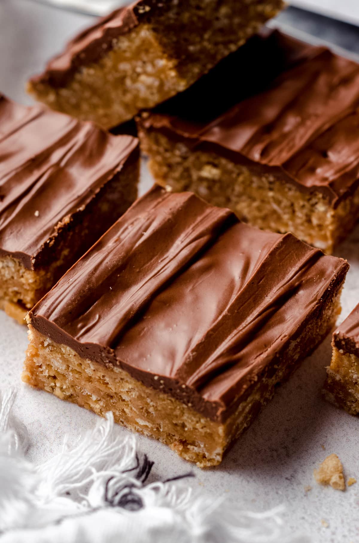 Scotcharoos cut into bars and sitting on a white counter. The base is a golden caramel color and it is topped with a layer of chocolate. 