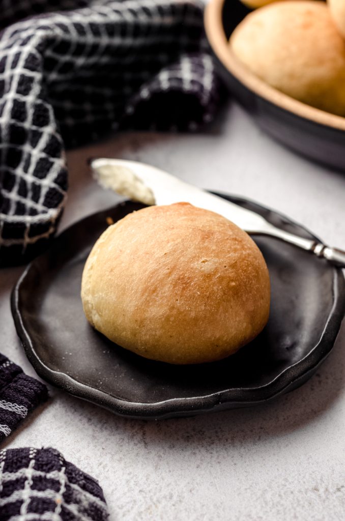 A sourdough dinner roll sitting on a black plate with a butter knife behind it.