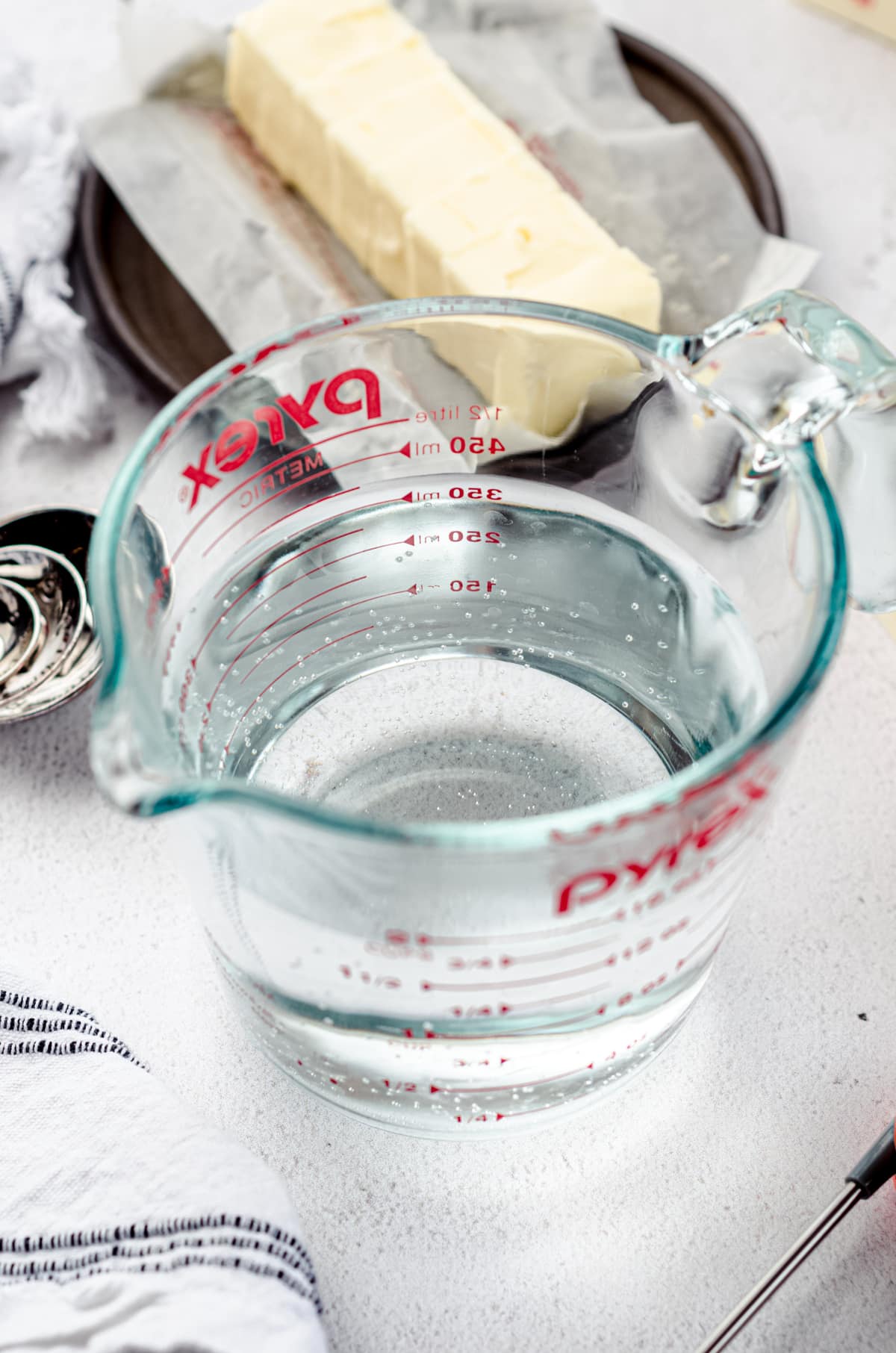 A glass measuring cup full of water with a stick of butter and measuring spoons in the background.