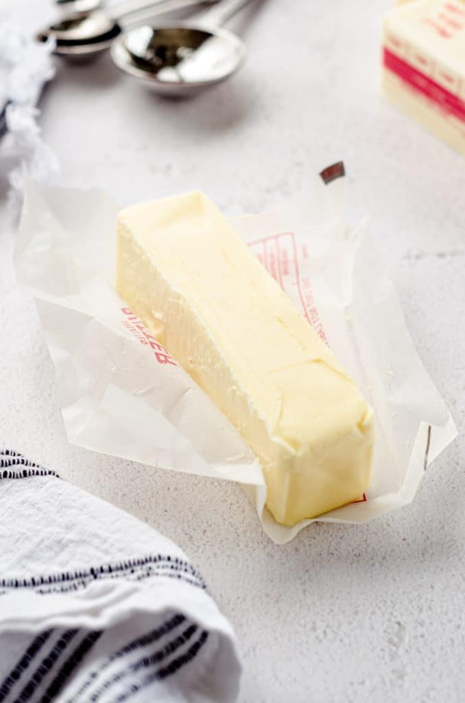 A stick of butter on its wrapper with measuring spoons in the background.