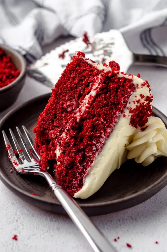 A slice of red velvet cake on a plate with a bite taken out of it and a fork resting on the plate.