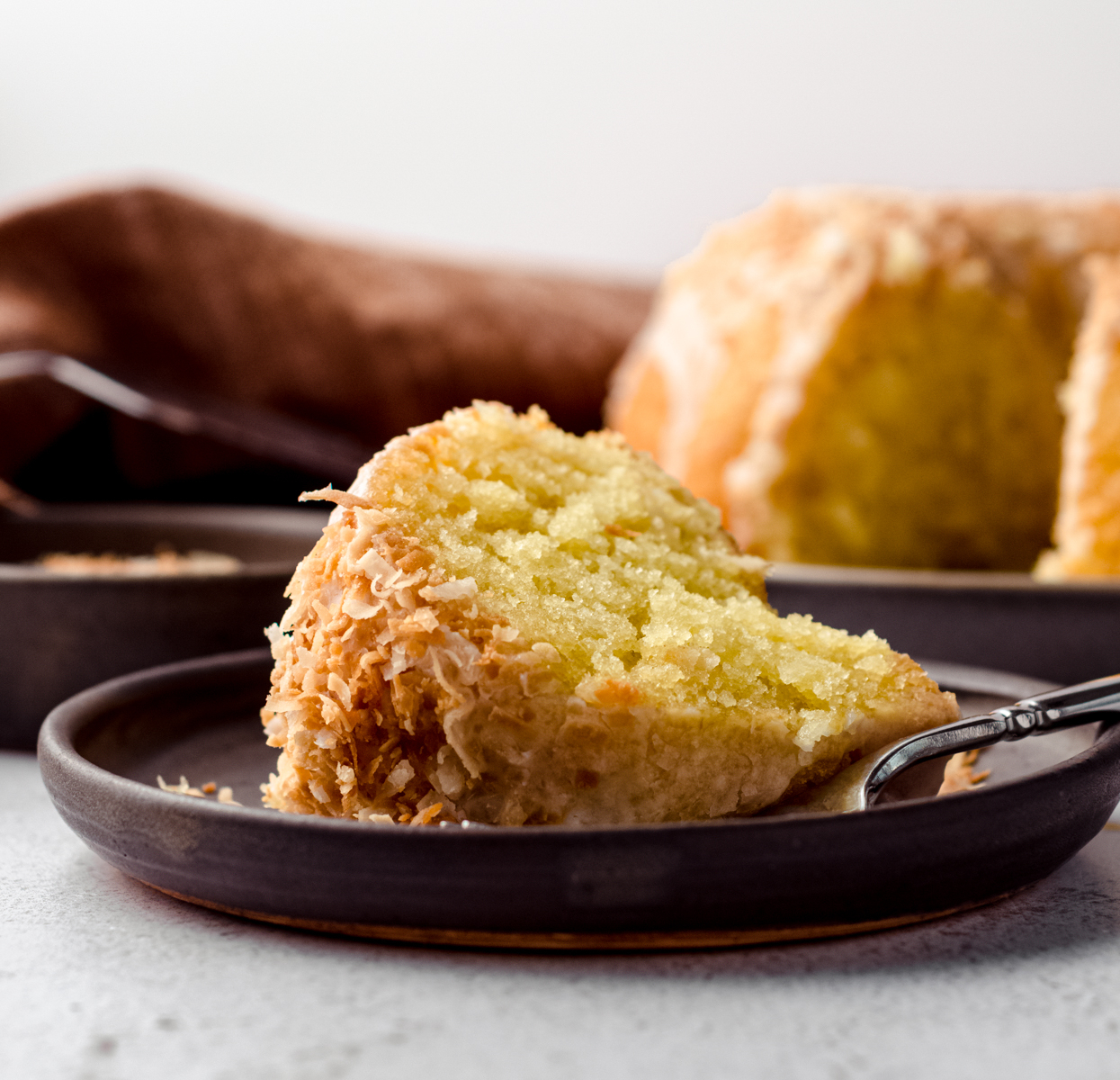 A slice of coconut bundt cake sitting on a plate with a fork. There is a coconut bundt cake in the background. 