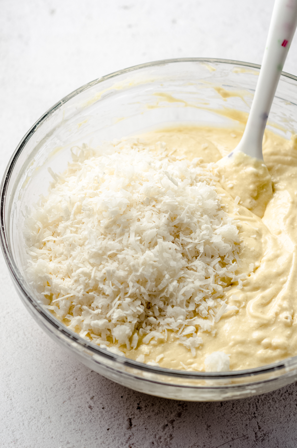 Glass mixing bowl with cake batter and shredded coconut with rubber spatula. 