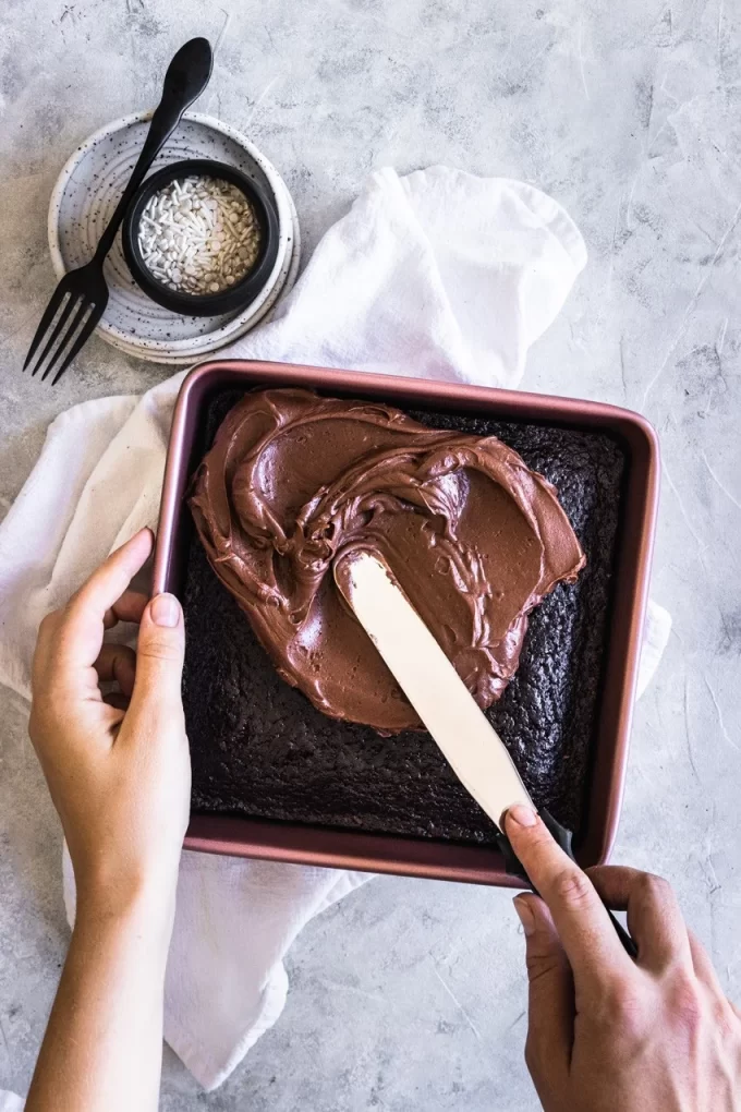 Aerial photo of a hand spreading chocolate frosting onto a sourdough chocolate cake in a pan.