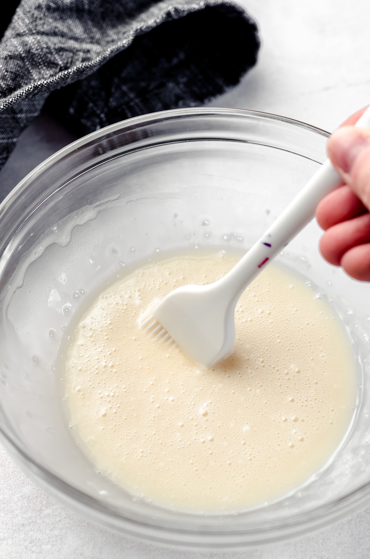 A hand holding a pastry brush in a bowl of vanilla glaze for hot cross buns.