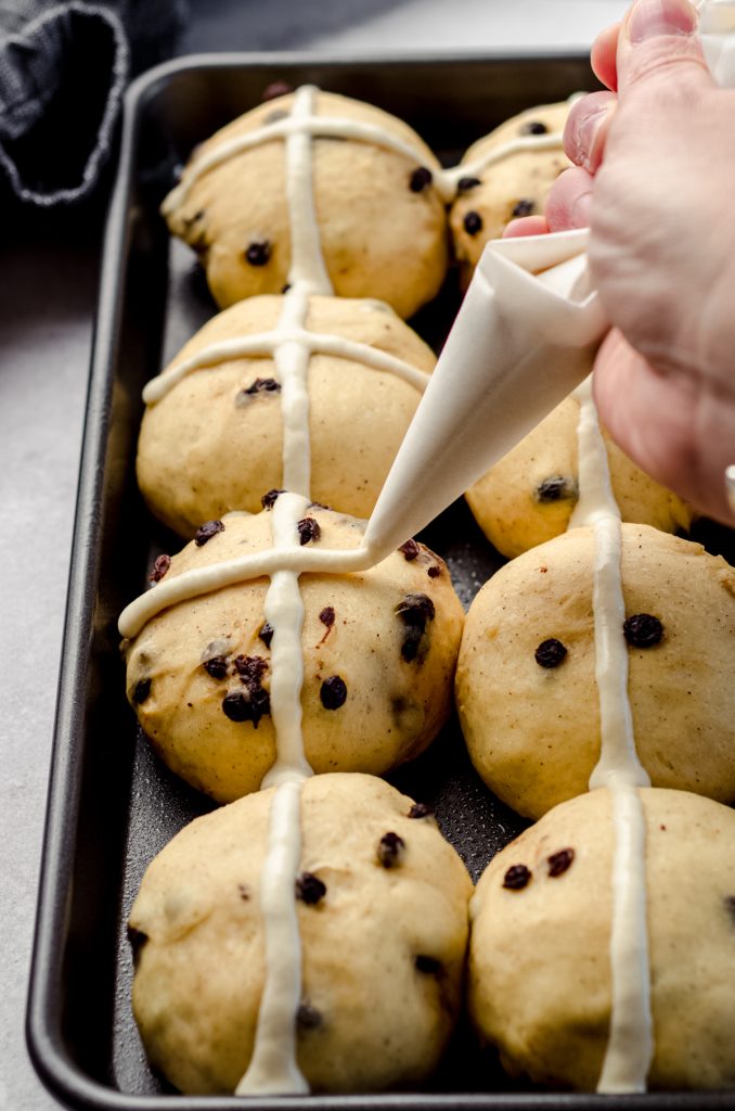 A hand piping a flour cross on top of hot cross buns.