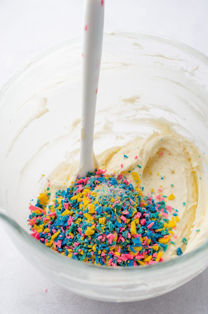 White chocolate rainbow chips in a bowl of vanilla frosting to make homemade rainbow chip frosting.