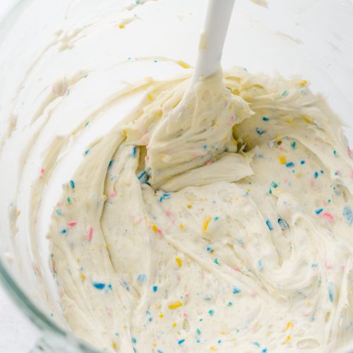 Rainbow chip frosting in the glass bowl of a stand mixer with a spatula.