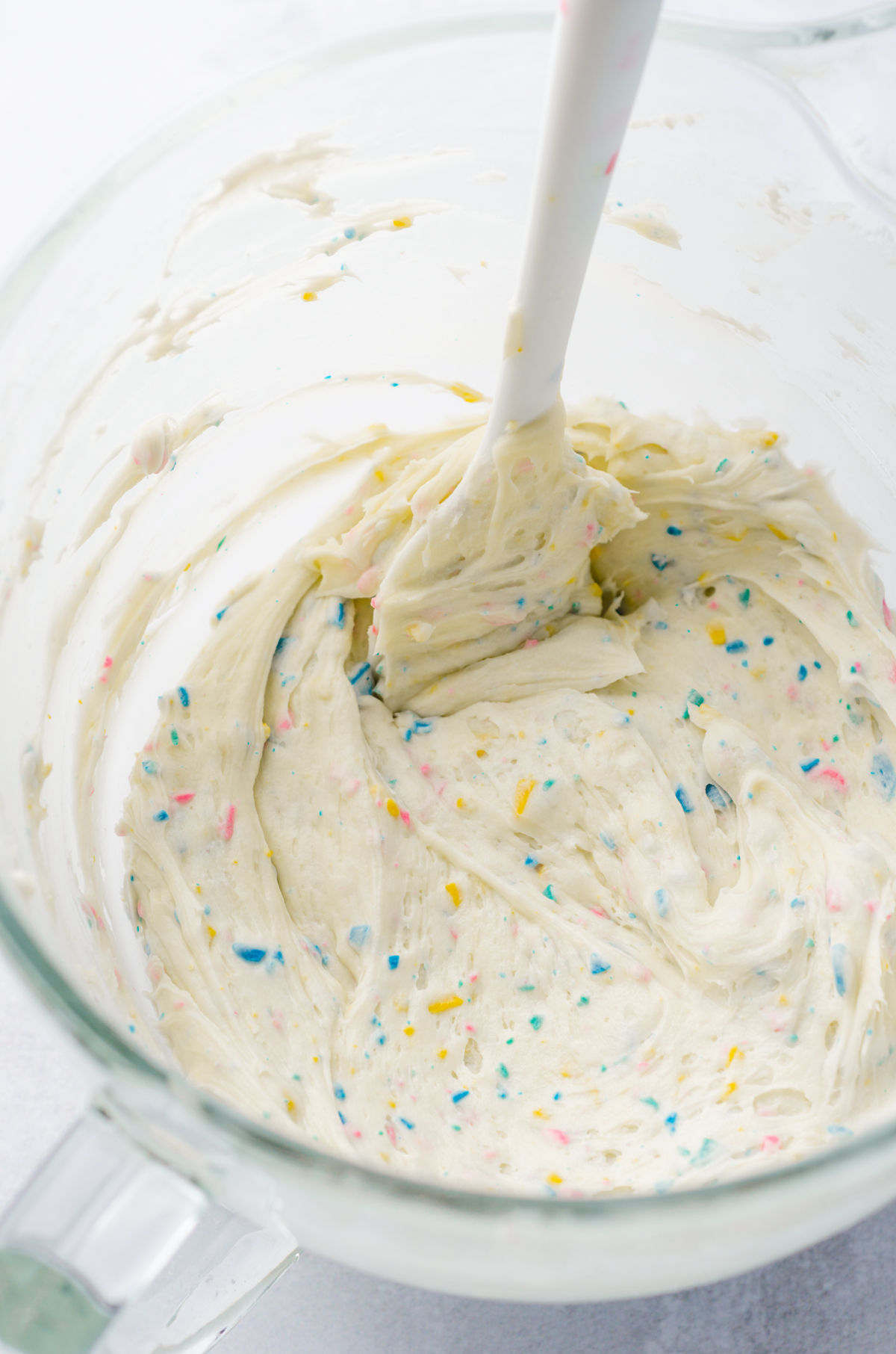 Rainbow chip frosting in the glass bowl of a stand mixer with a spatula.