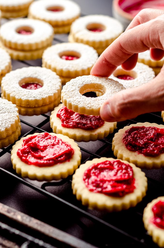 A hand placing the top cookie on a Linzer cookie filled with raspberry filling.