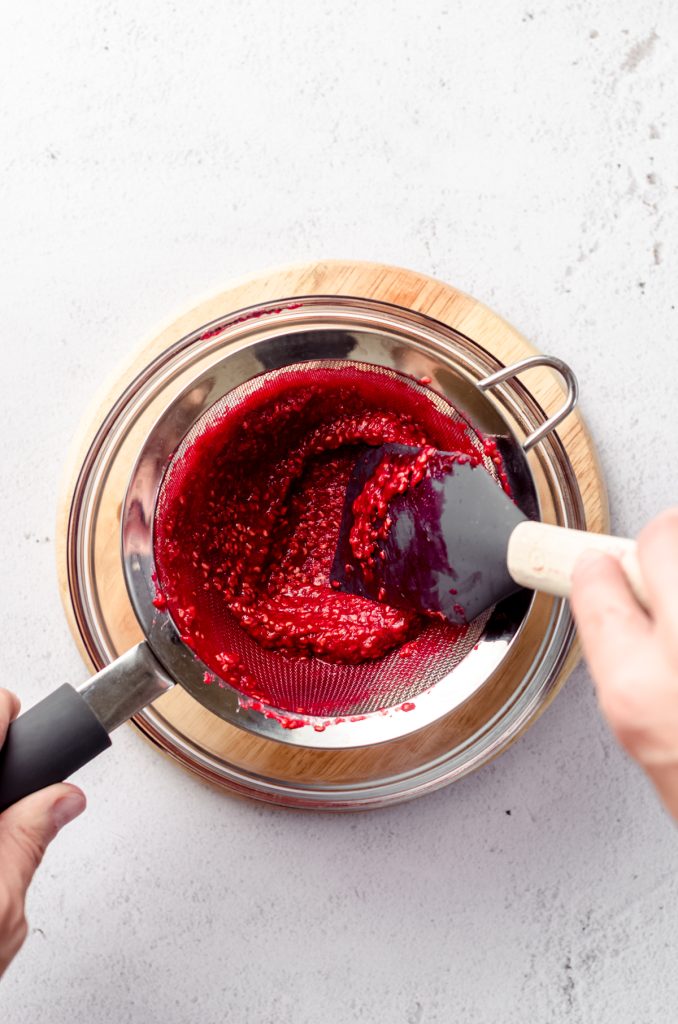 Someone is pressing cooked raspberry spread through a mesh sieve to remove seeds.
