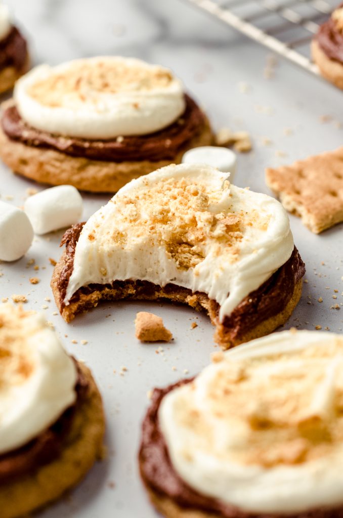 Frosted s'mores cookies on a surface with graham crackers and marshmallows scattered around. There is a bite taken out of the one in the front.