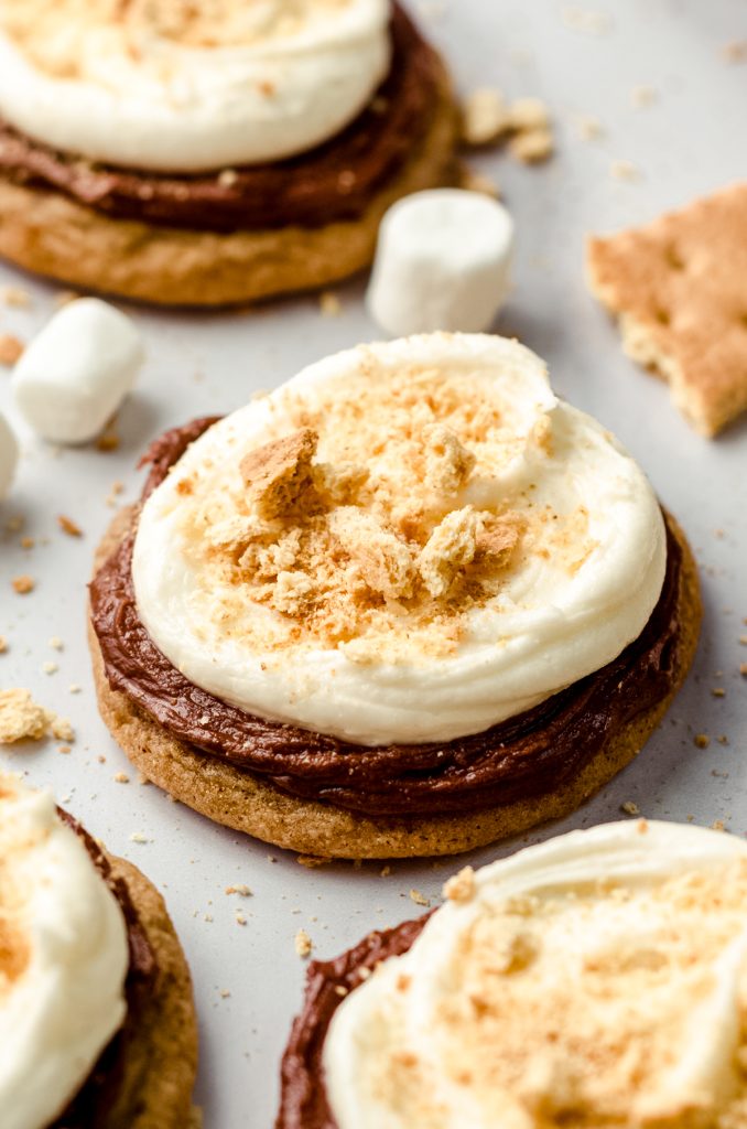 Frosted s'mores cookies on a surface with graham crackers and marshmallows scattered around.