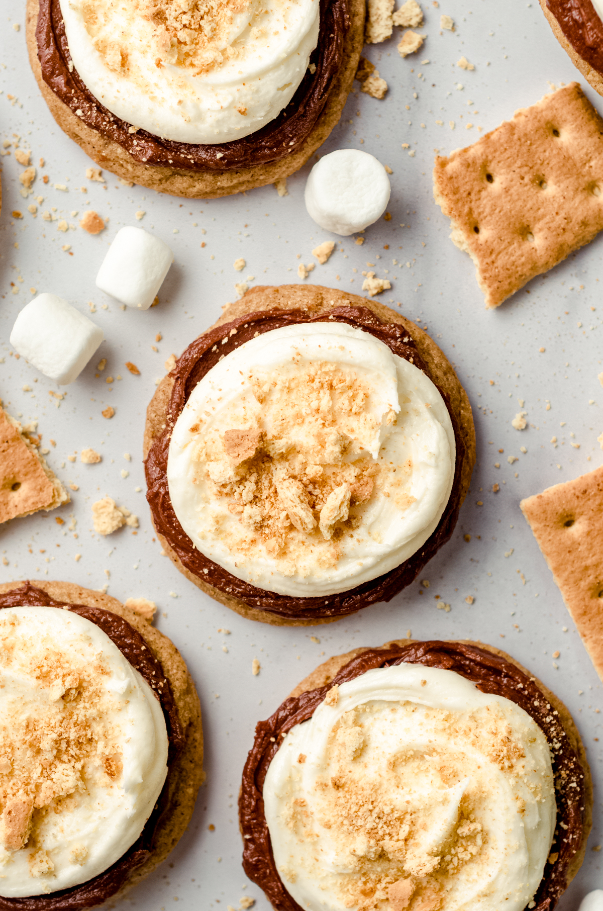 Aerial photo of frosted s'mores cookies on a surface with graham crackers and marshmallows scattered around.