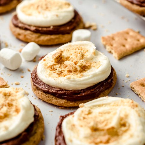 Frosted s'mores cookies on a surface with graham crackers and marshmallows scattered around.