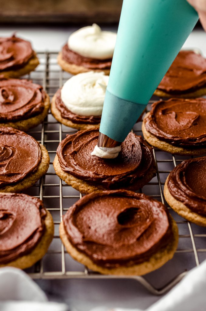 Graham cracker cookies with chocolate frosting spread on top and someone piping marshmallow frosting on top of that to make frosted s'mores cookies.