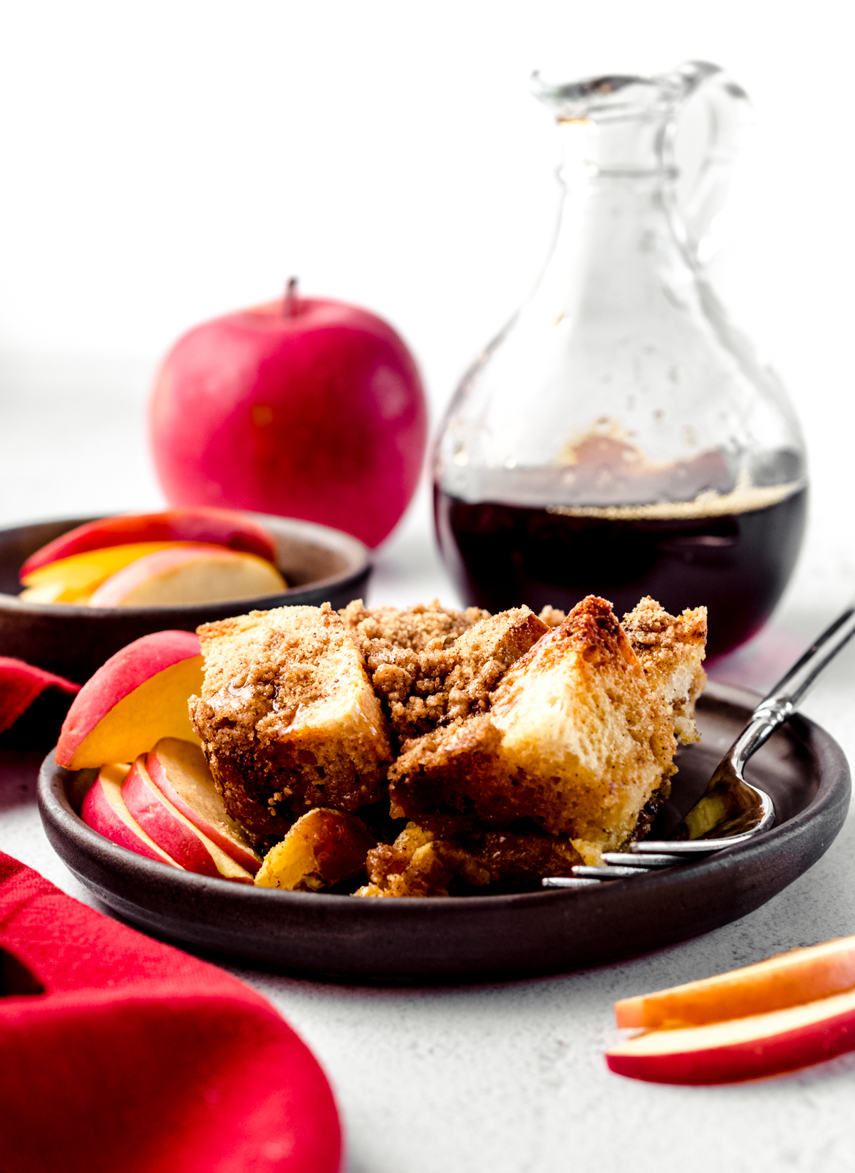 A serving of apple French toast casserole on a dish with a fork and a container of syrup and an apple in the background.