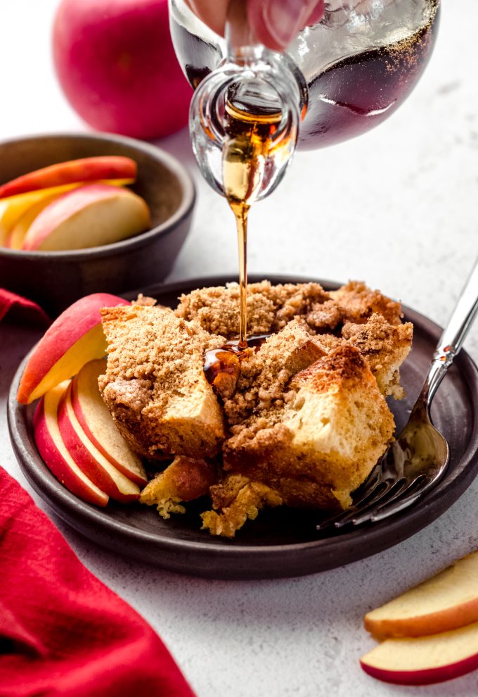 Someone is pouring syrup onto a serving of apple French toast casserole on a dish with a fork.