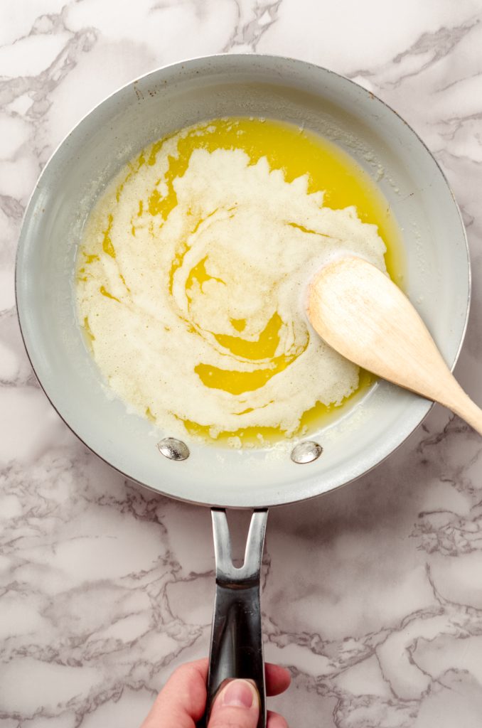 An aerial photo of a skillet of melting butter to make brown butter. There is a hand holding the handle of the skillet and a wooden spoon stirring the butter.