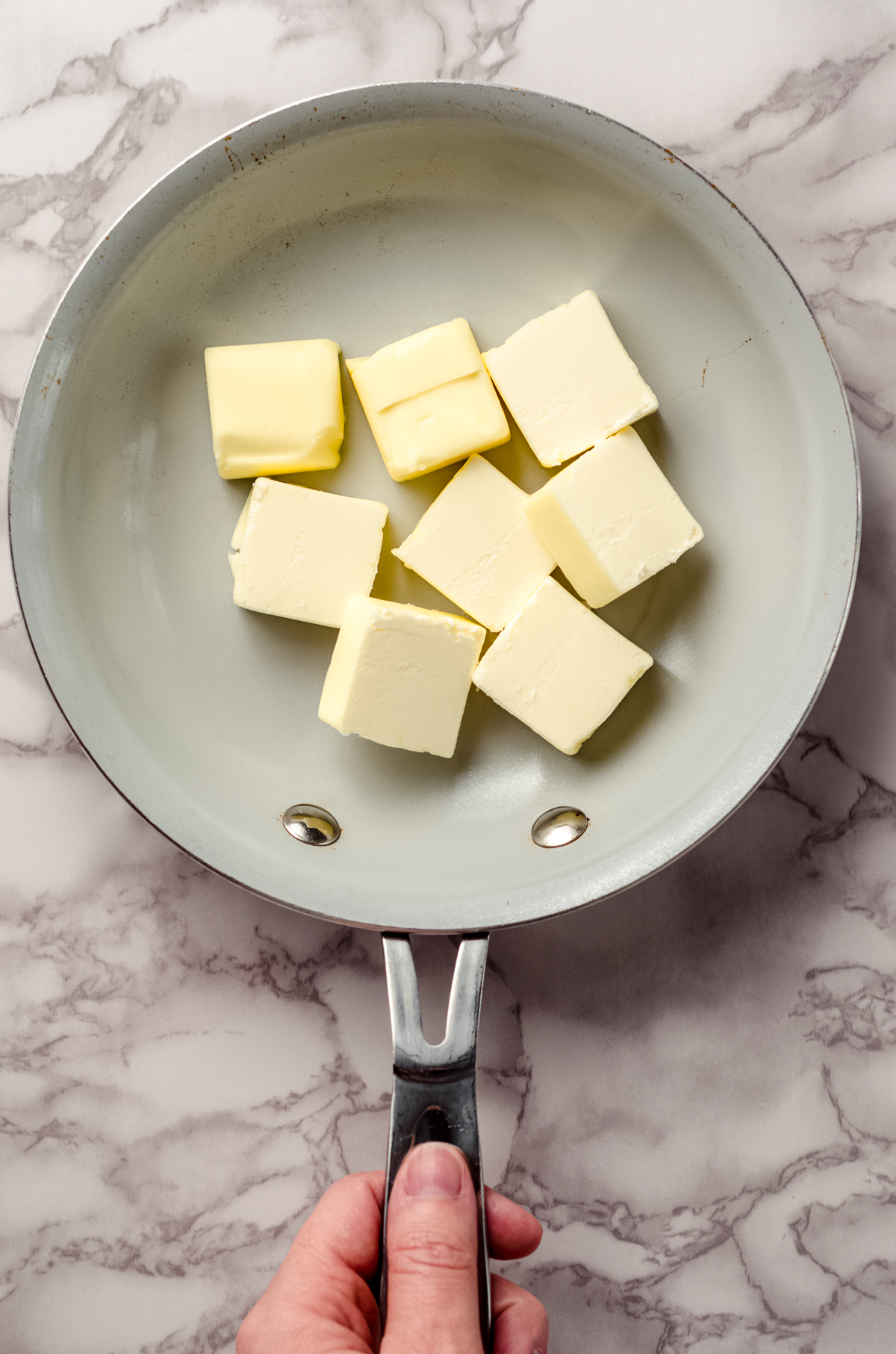 An aerial photo of a skillet of butter ready to make brown butter. There is a hand holding the handle of the skillet.