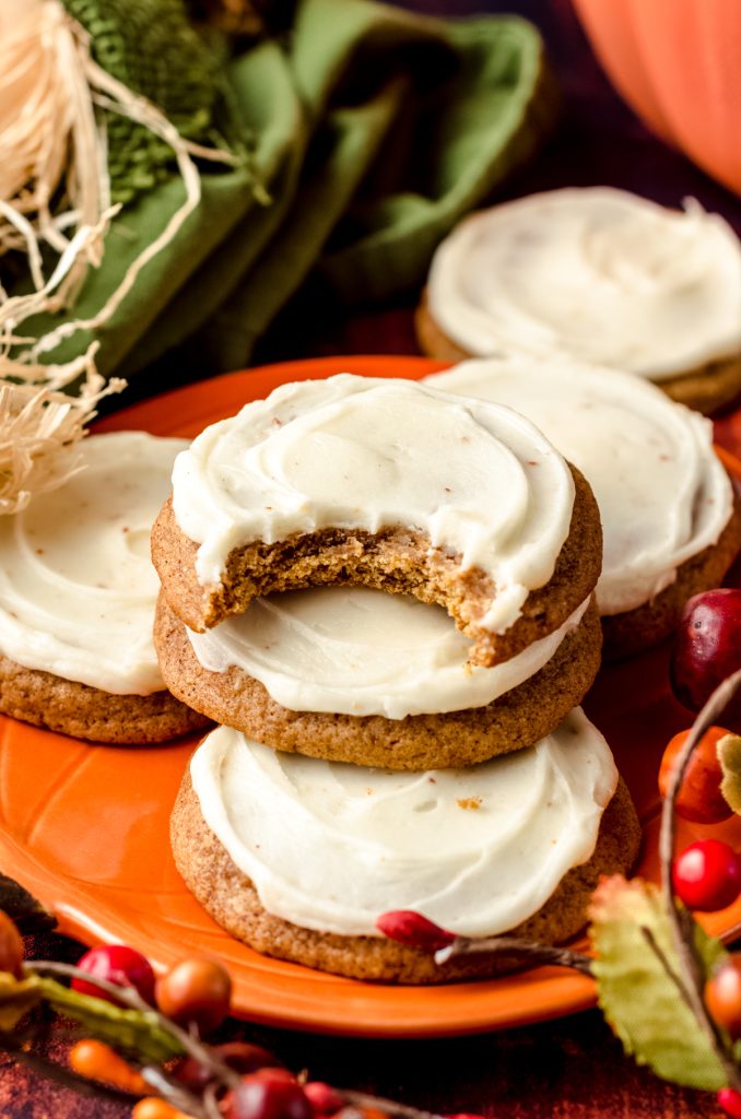 A stack of frosted pumpkin cookies on a plate with fall decor all around. There is a bite taken out of the cookie on top.