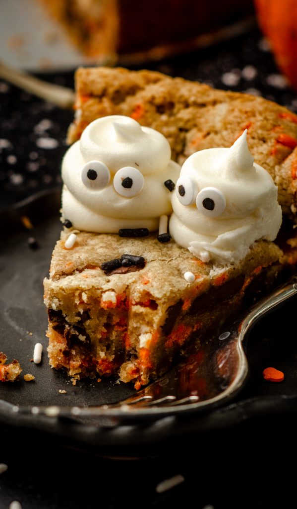 A slice of Halloween cookie cake with buttercream ghosts piped onto it on a black plate with a fork. A bite has been taken out of the slice.