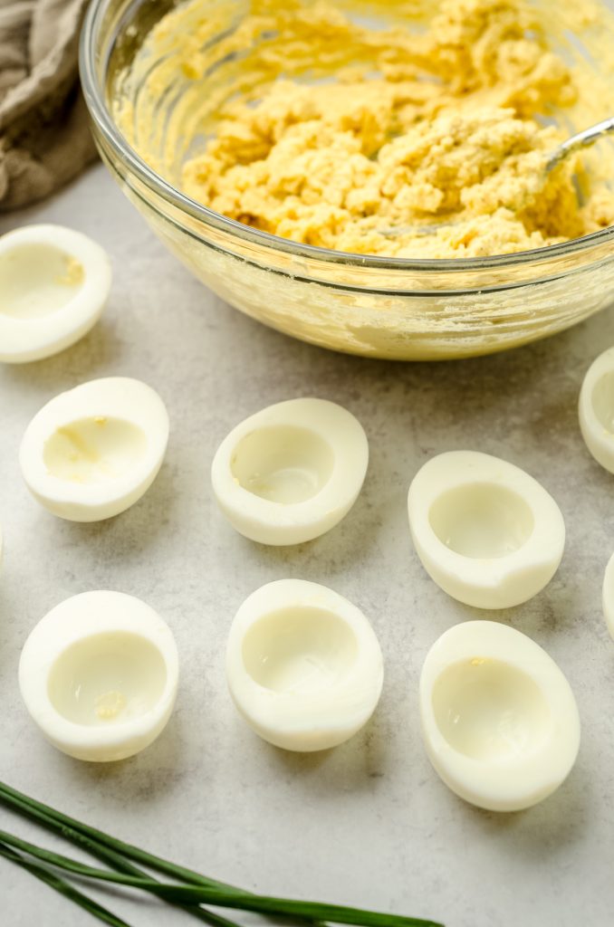 Halved cooked egg whites with a bowl of filling behind them to make deviled eggs.