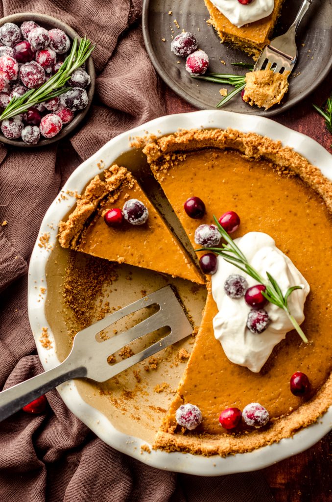 An aerial photo of pumpkin pie with graham cracker crust sliced in a pie plate and ready to serve. There is whipped cream, cranberries, and fresh rosemary garnish on the pie.