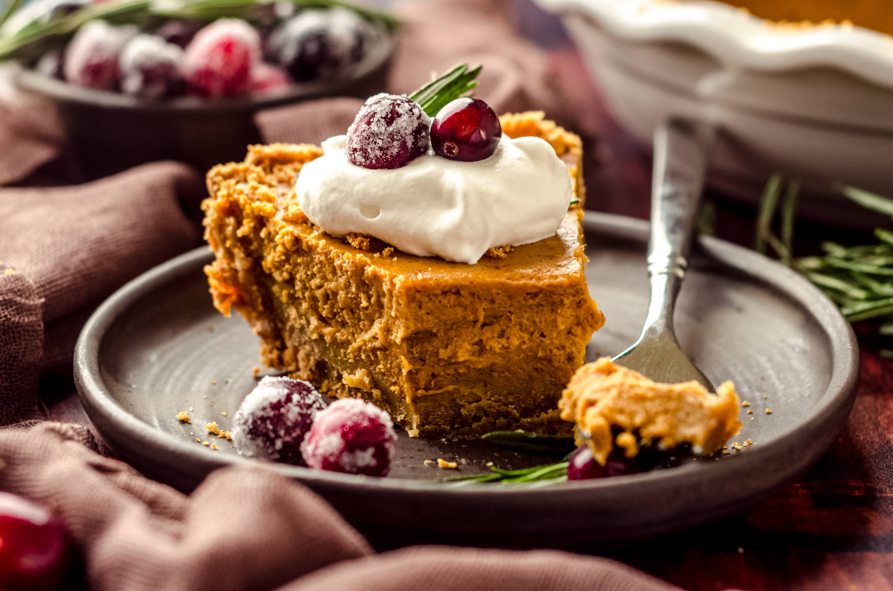A slice of pumpkin pie with graham cracker crust on a plate with a dollop of whipped cream and cranberries and rosemary garnishing the slice. A bite has been taken out of the pie with a fork.