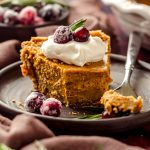 A slice of pumpkin pie with graham cracker crust on a plate with a dollop of whipped cream and cranberries and rosemary garnishing the slice. A bite has been taken out of the pie with a fork.