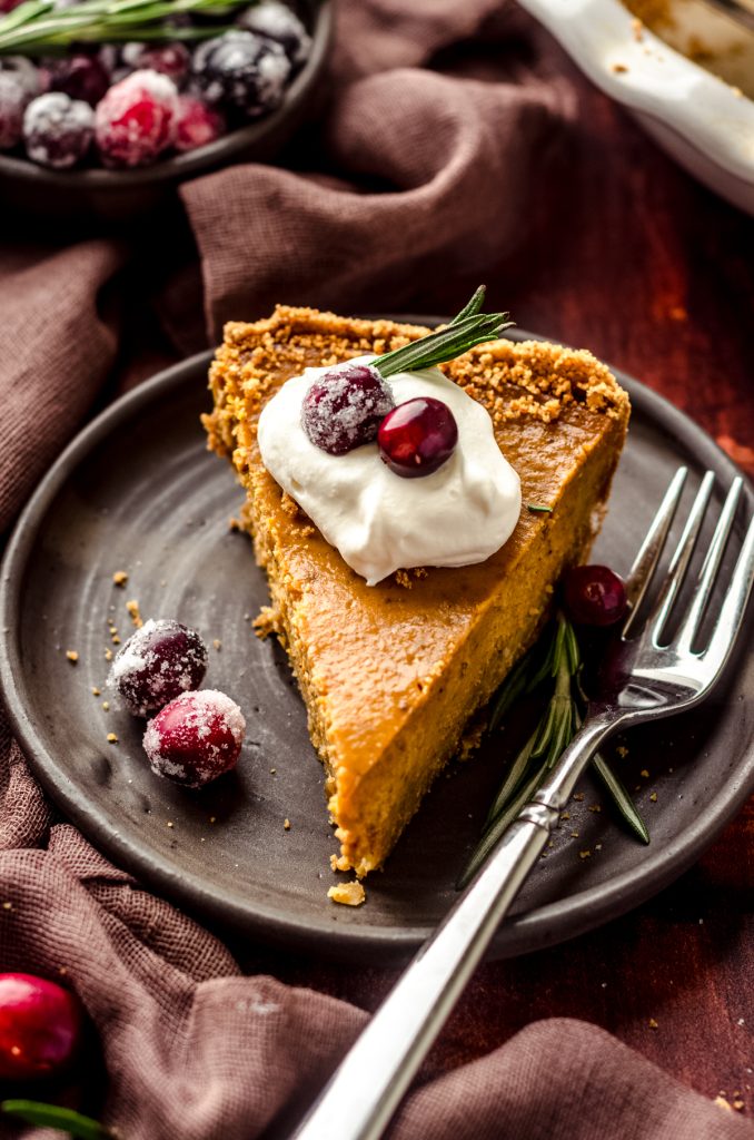 A slice of pumpkin pie with graham cracker crust on a plate with a dollop of whipped cream and cranberries and rosemary garnishing the slice.