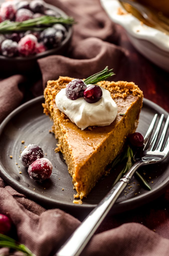 A slice of pumpkin pie with graham cracker crust on a plate with a dollop of whipped cream and cranberries and rosemary garnishing the slice.