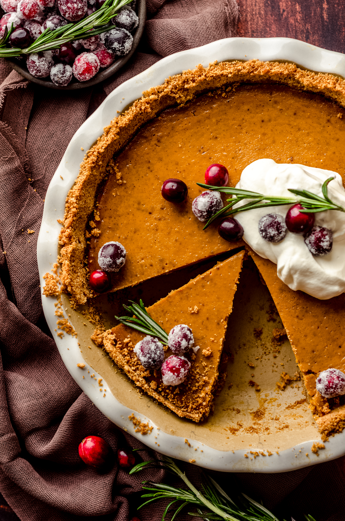 An aerial photo of pumpkin pie with graham cracker crust sliced in a pie plate and ready to serve. There is whipped cream, cranberries, and fresh rosemary garnish on the pie.