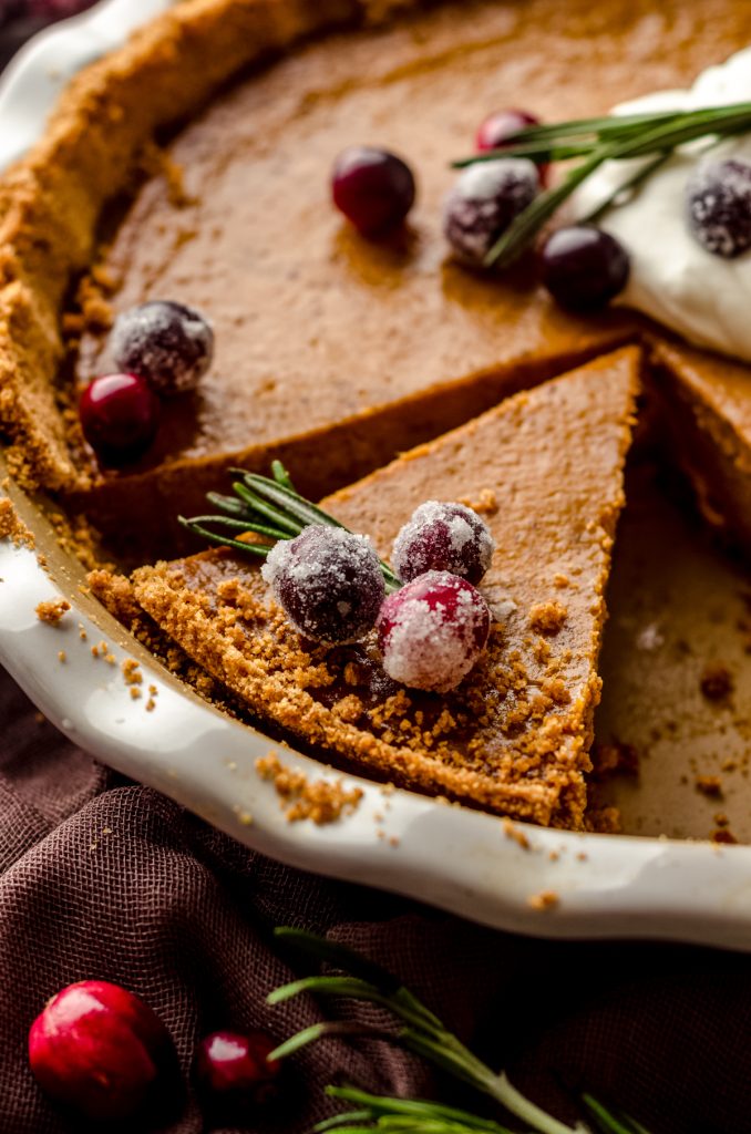 Pumpkin pie with graham cracker crust sliced in a pie plate and ready to serve. There is whipped cream, cranberries, and fresh rosemary garnish on the pie.