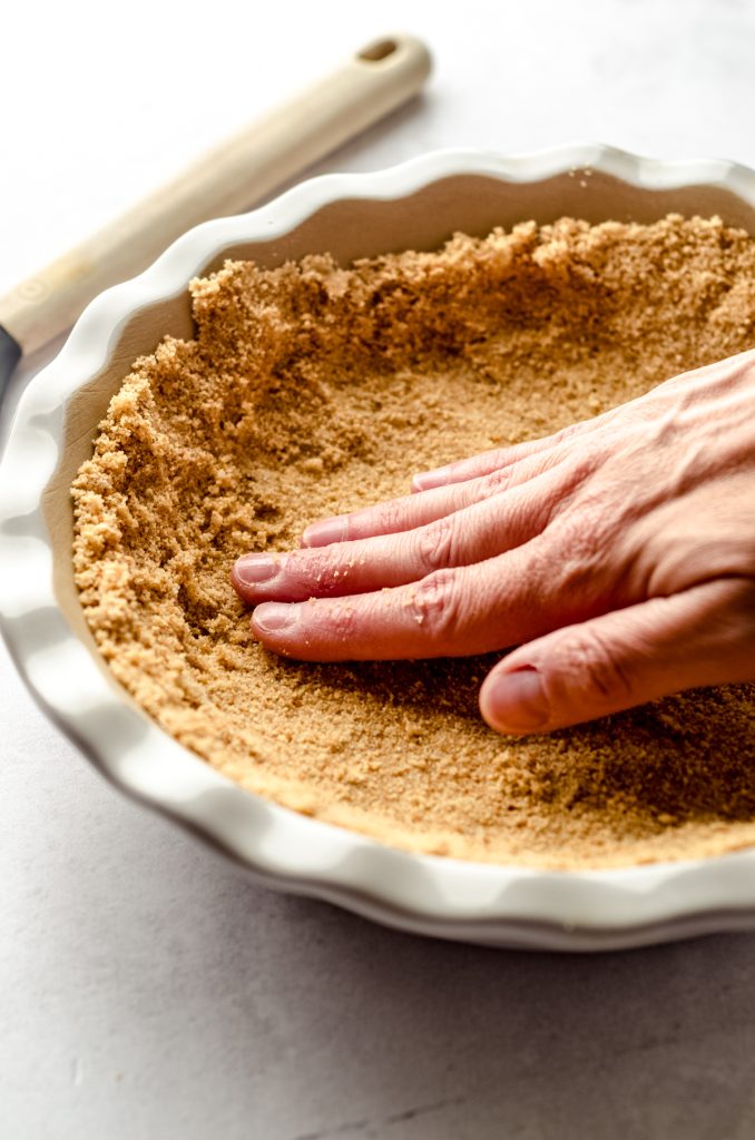 Someone is using their hand to press graham cracker crust mixture into a pie plate.