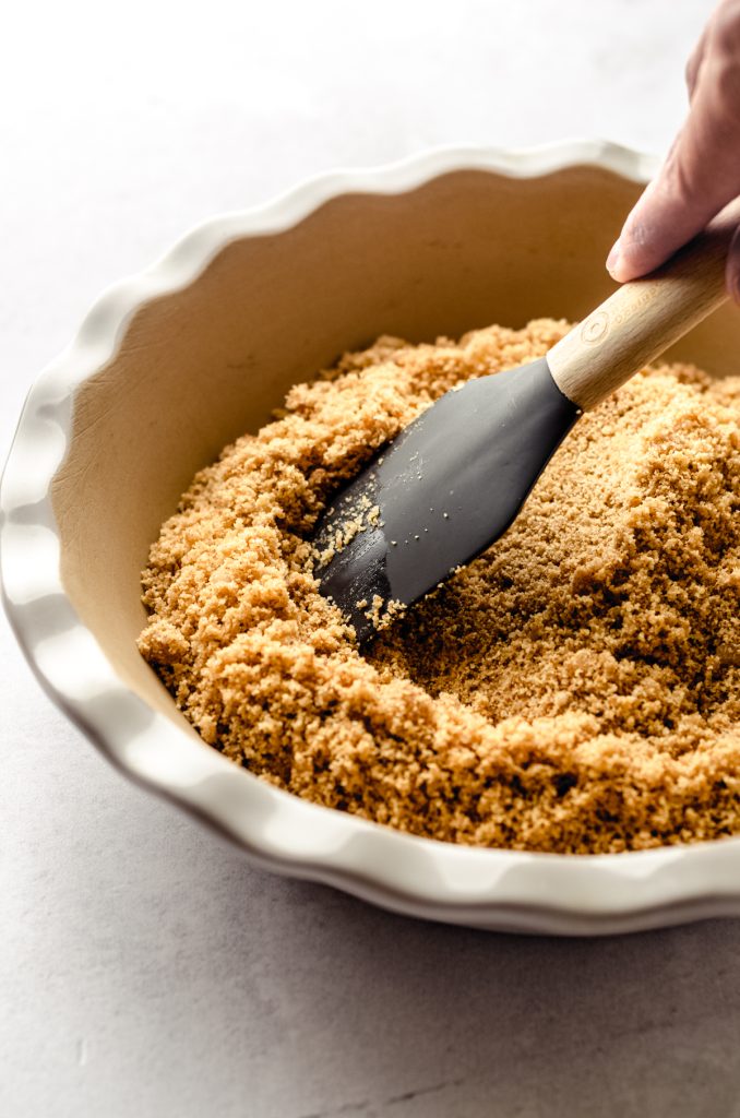 Someone is using a spatula to press graham cracker crust mixture into a pie plate.