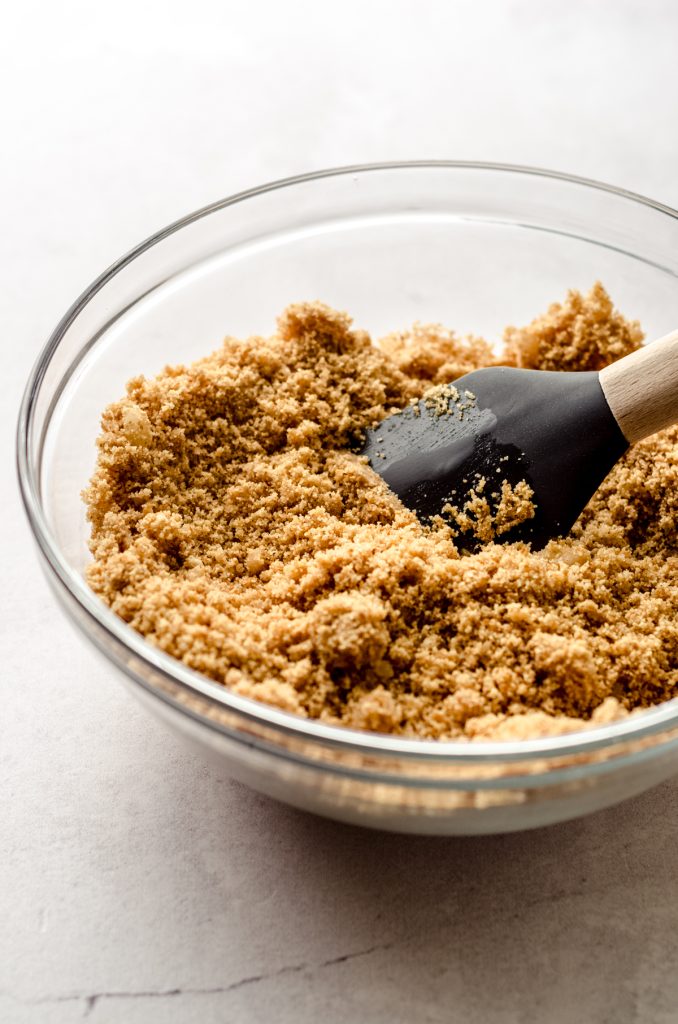 Someone is stirring a bowl of ingredients for graham cracker crust with a spatula.