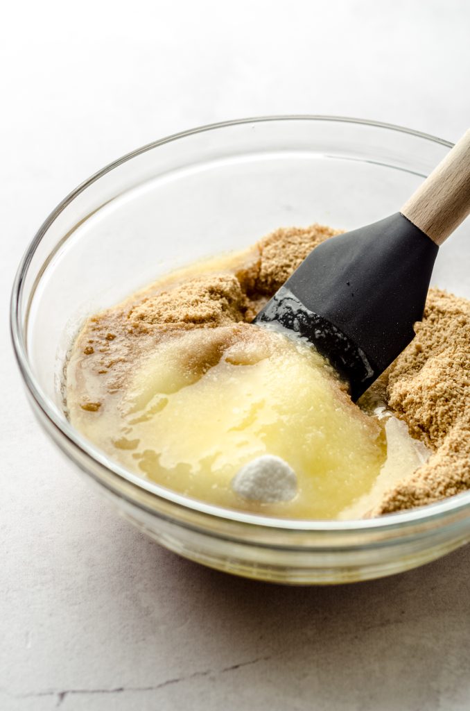Someone is stirring a bowl of ingredients for graham cracker crust with a spatula.