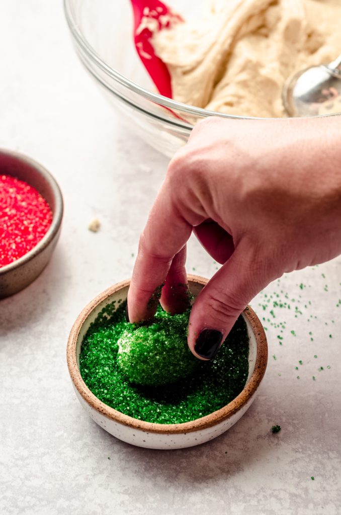 Someone is using their fingers to roll a ball of cookie dough into green sanding sugar.