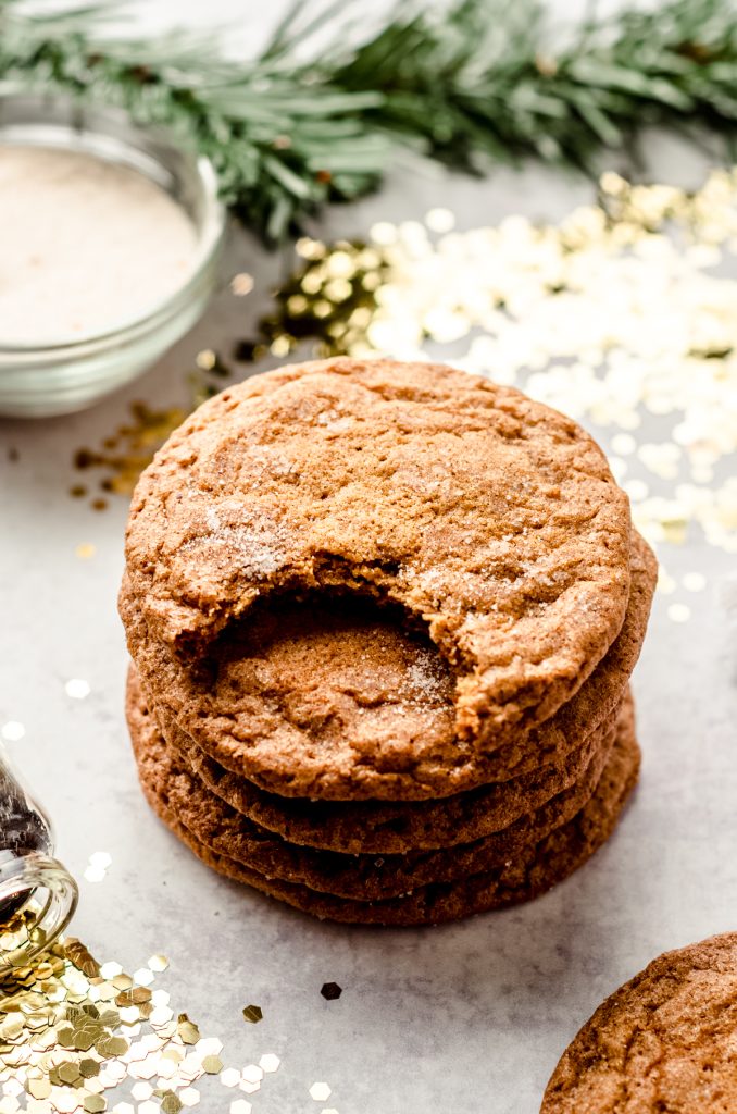 A stack of crispy gingersnap cookies on a plate with gold confetti, evergreen sprigs, and a bowl of spiced sugar around it. The cookie on top has a bite taken out of it.