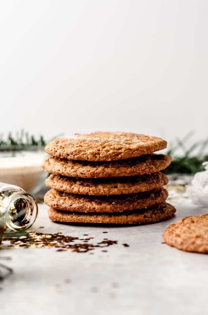 A stack of crispy gingersnap cookies on a plate with gold confetti, evergreen sprigs, and a bowl of spiced sugar around it.