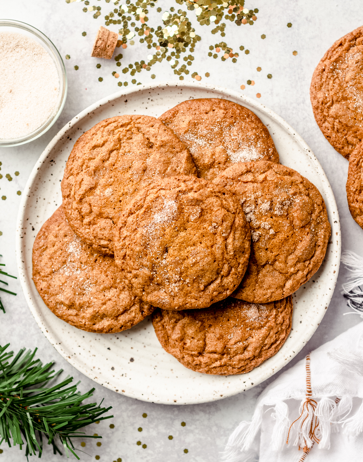 Aerial photo of crispy gingersnap cookies on a plate with gold confetti, evergreen sprigs, and a bowl of spiced sugar around it.